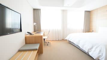 Una habitación de hotel moderna con una cama grande, un escritorio con silla, un televisor montado en la pared y un amplio ventanal con cortinas.