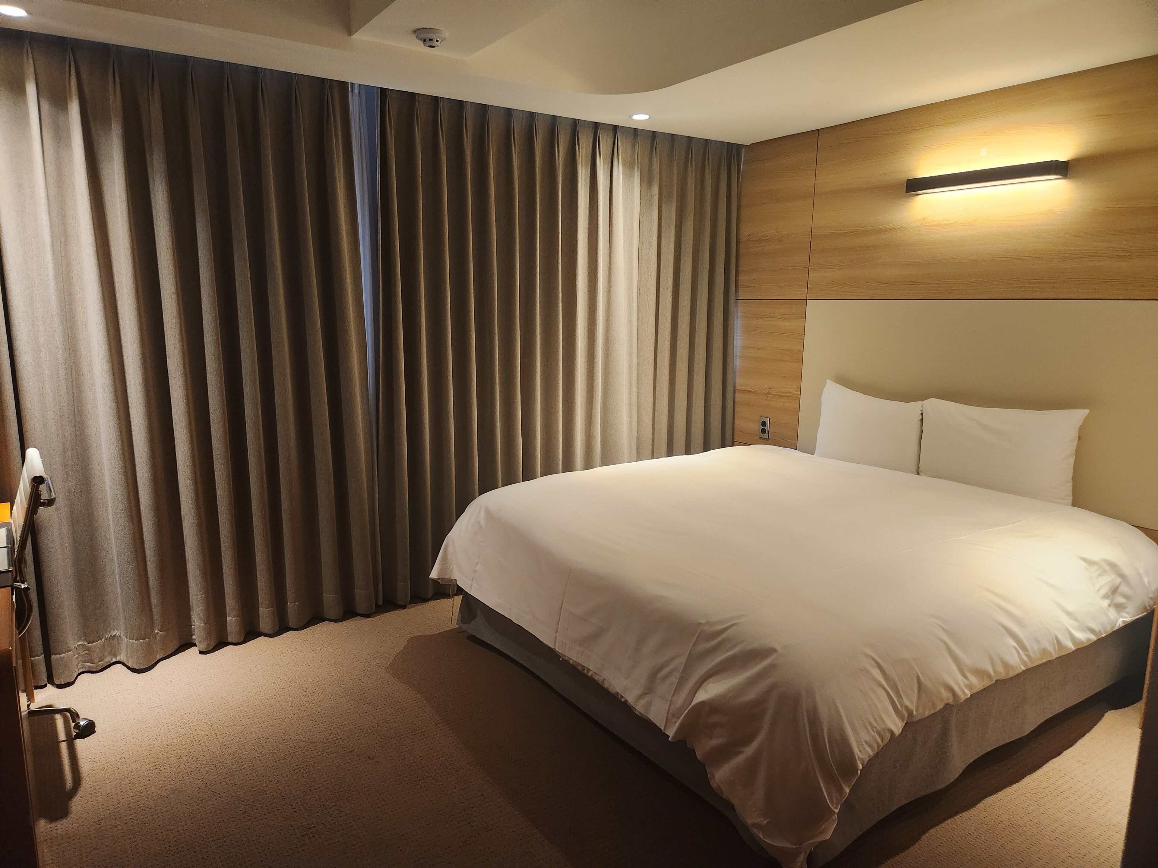 Een hotelkamer met een groot bed, beige gordijnen, een houten hoofdbord en een wandlamp.