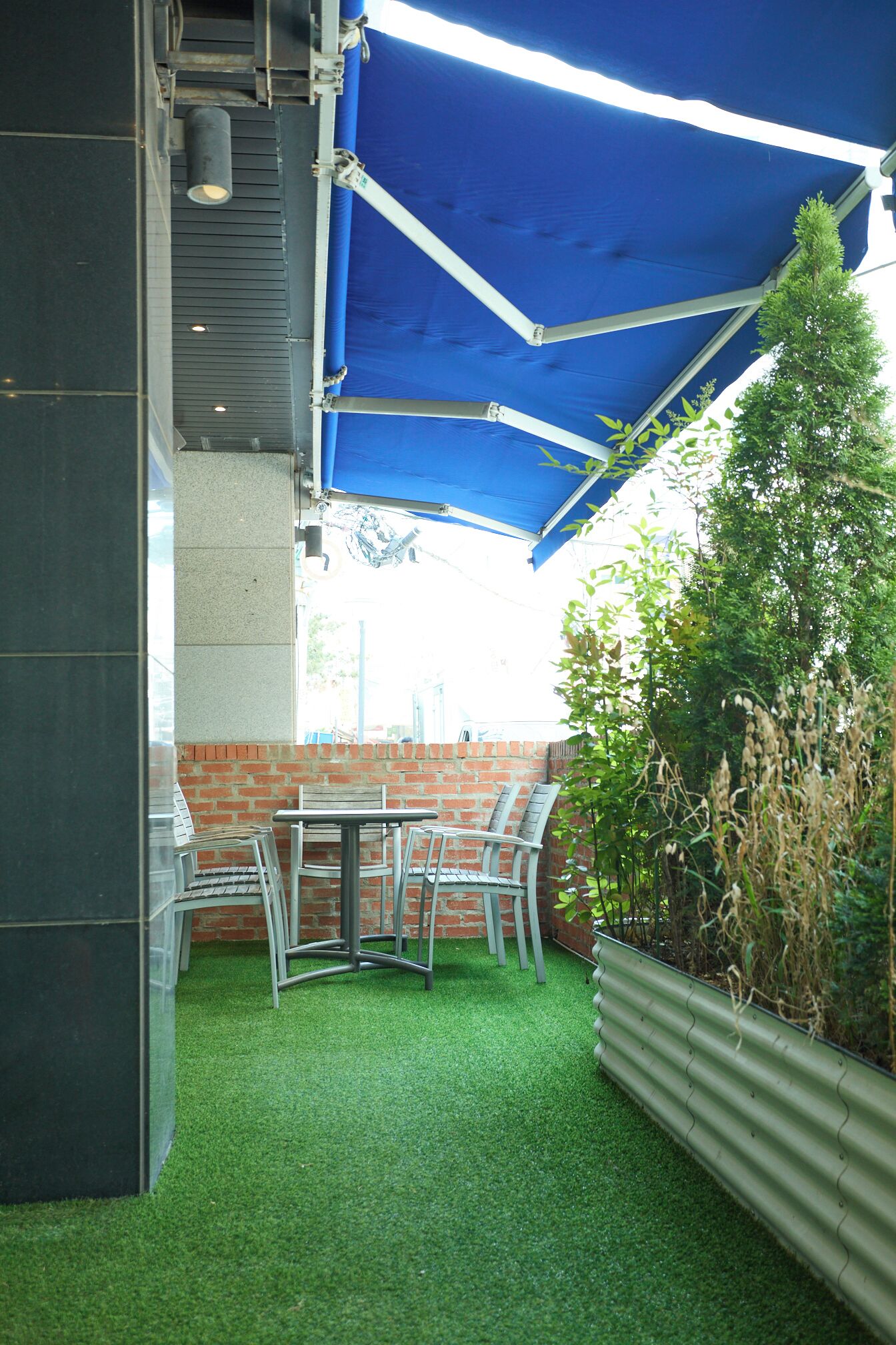 A patio area with a table and chairs, artificial grass, a brick wall, and a blue canopy.