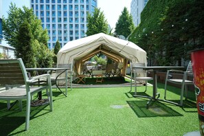 A tented outdoor area with artificial grass, surrounded by chairs and tables, and buildings in the background. - Acube Hotel (Seoul)