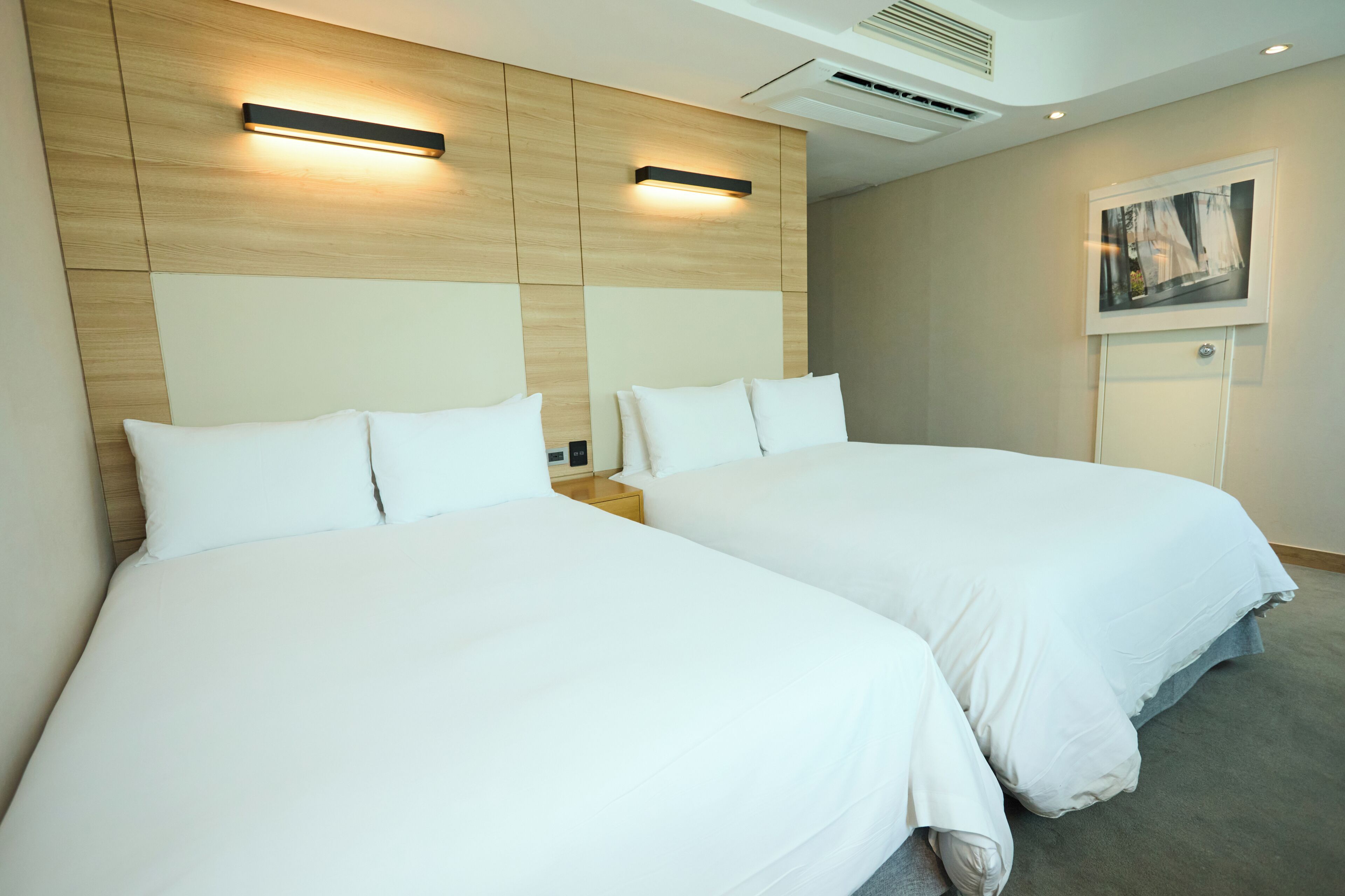 A hotel room with two beds, a wooden headboard, a nightstand, a framed picture on the wall, and an air conditioning unit.