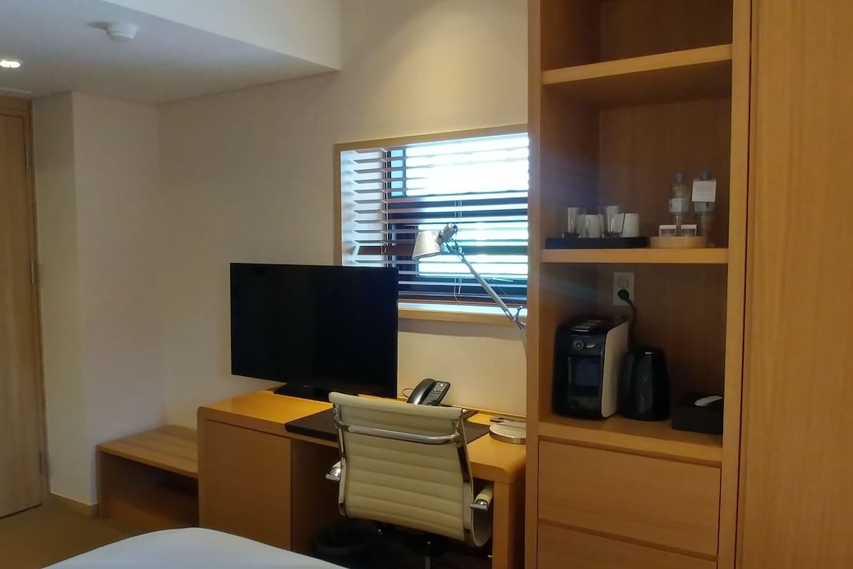 A hotel room with a bed, a desk with a chair, a television, a window with blinds, and a shelf with toiletries.