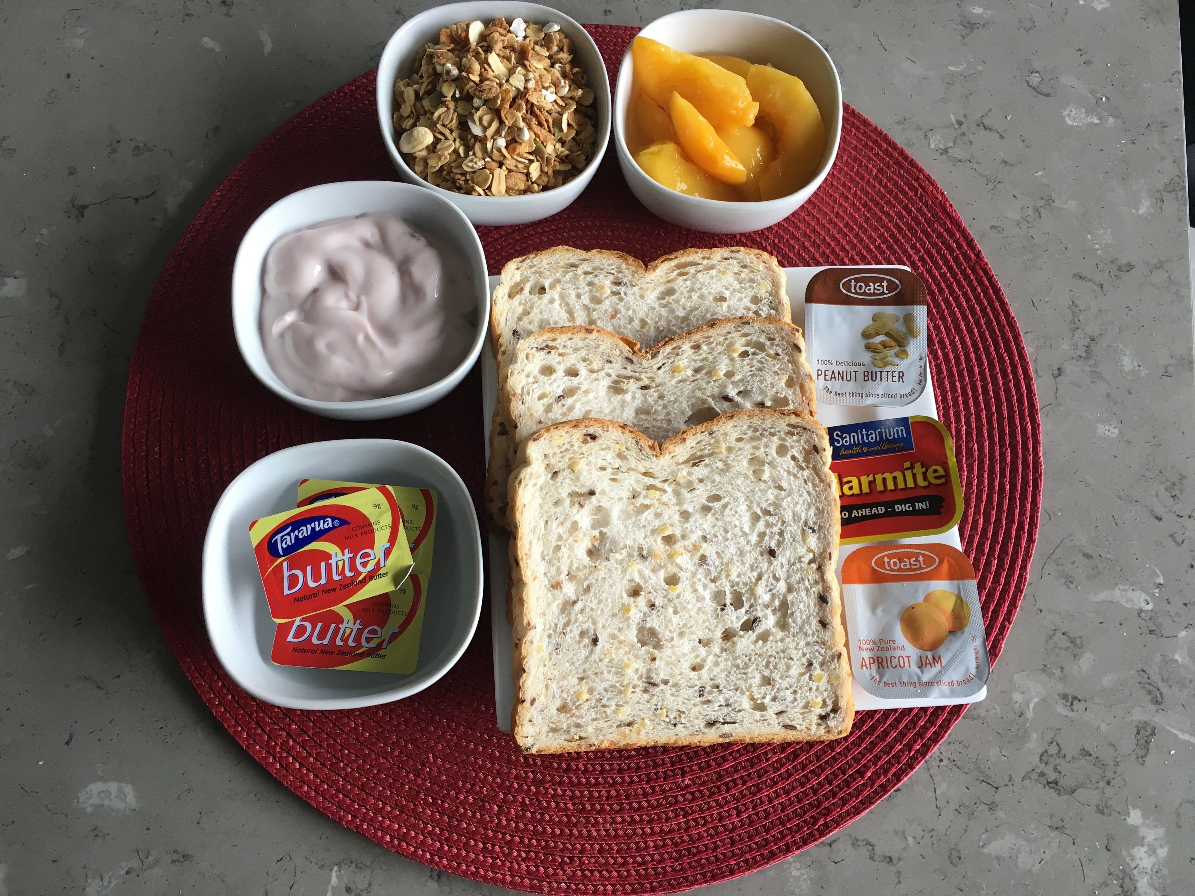 Desayuno con tostadas, mantequilla de maní, mermelada, yogur, granola y melocotones.