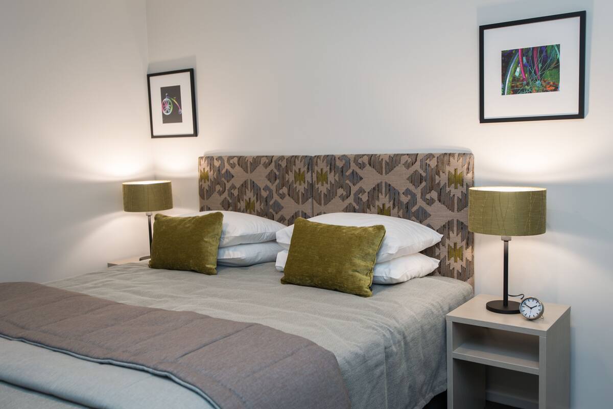 A neatly made bed with two pillows, a bedside table with a clock, and two wall-mounted lamps.