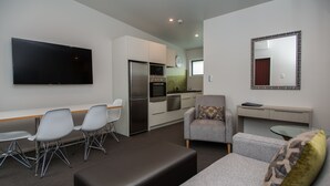 A modern living room with a flat-screen TV, grey sofas, a dining area with chairs, a kitchen with appliances, and a mirror on the wall. - Pedlars Motel (Paeroa)