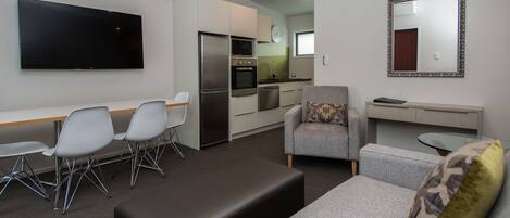 A modern living room with a flat-screen TV, grey sofas, a dining area with chairs, a kitchen with appliances, and a mirror on the wall.