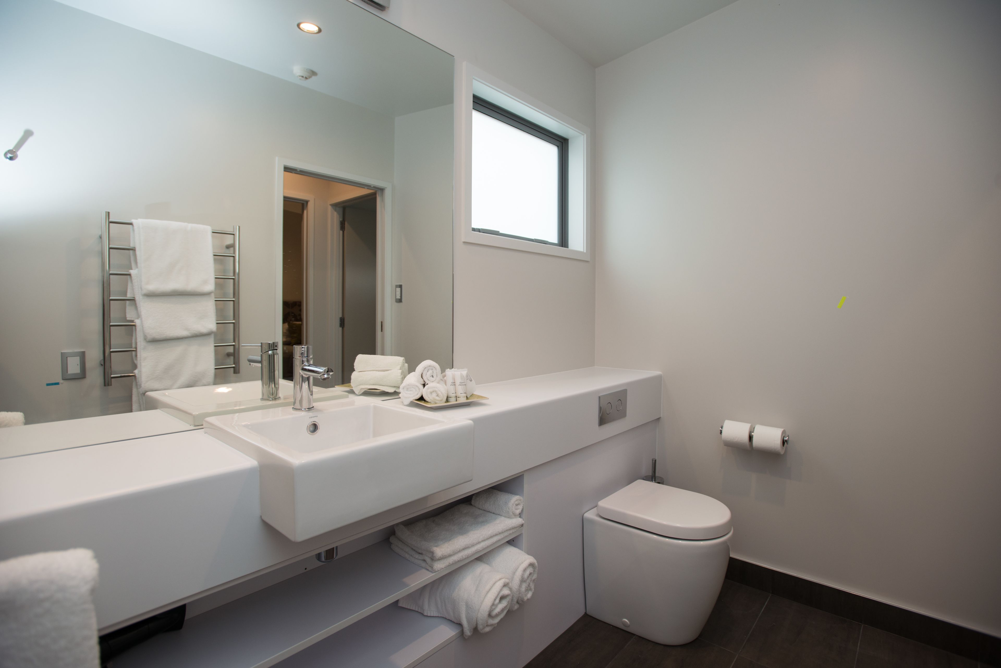 A modern bathroom with a large mirror, two sinks, a toilet, and a towel rack.