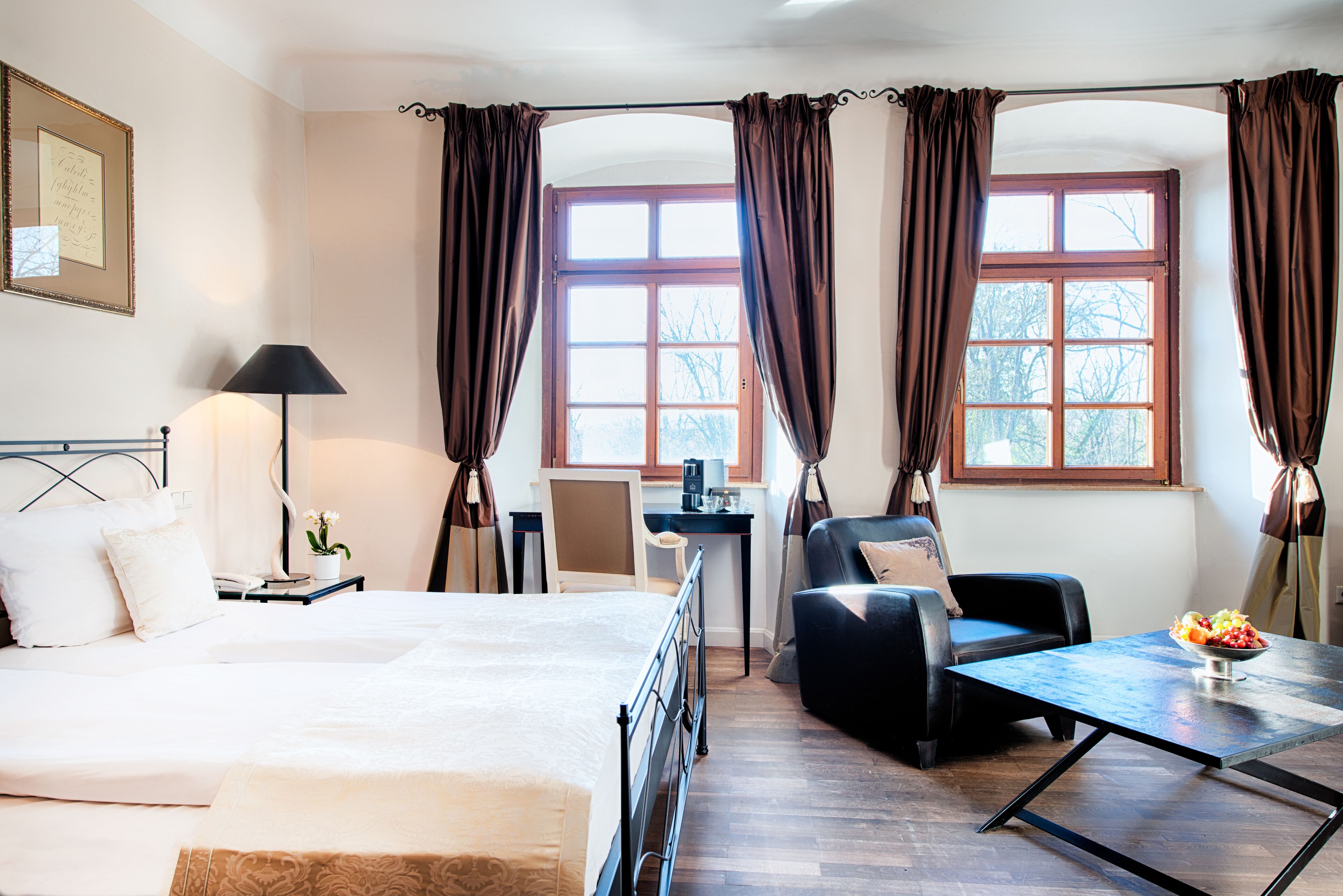 Une chambre d’hôtel avec un lit, un bureau, une chaise, une table avec un saladier de fruits et de grandes fenêtres avec des rideaux.