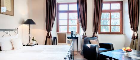 Une chambre d’hôtel avec un lit, un bureau, une chaise, une table avec un saladier de fruits et de grandes fenêtres avec des rideaux.
