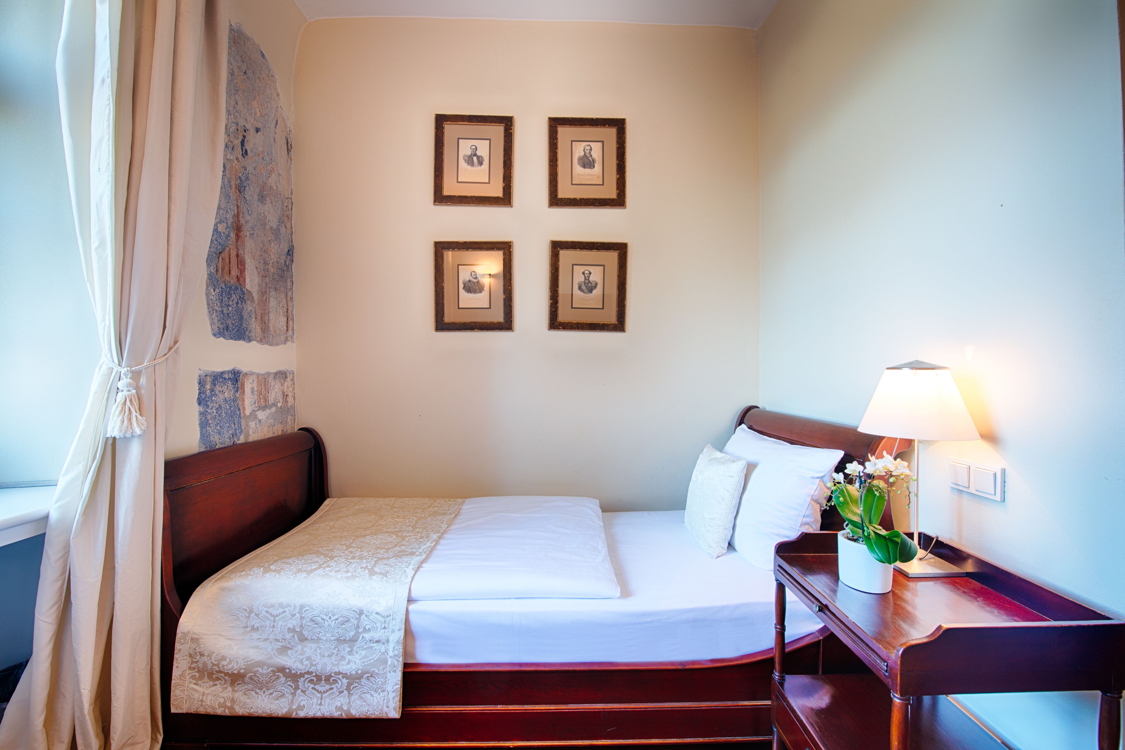 A neatly made bed with a white bedspread and pillows, a bedside table with a lamp and a plant, and four framed pictures on the wall.