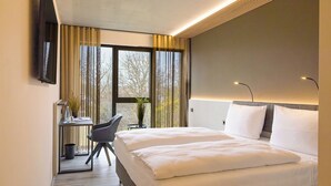 A hotel room with a bed, a desk with a chair, a TV mounted on the wall, and a window with curtains. - Hotel Schloss Lehen (Bad Friedrichshall)