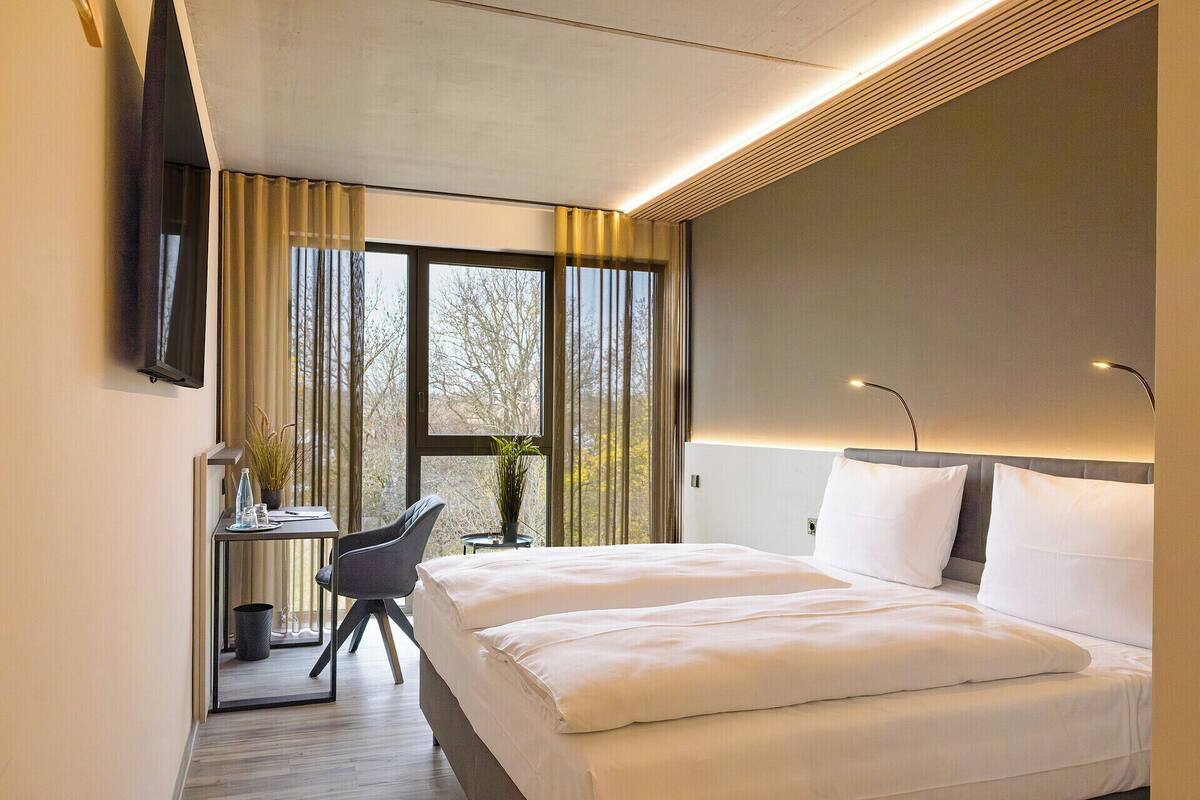 Habitació d'hotel amb un llit, una escriptori amb una cadira, un televisor fixat a la paret i una finestra amb cortines.