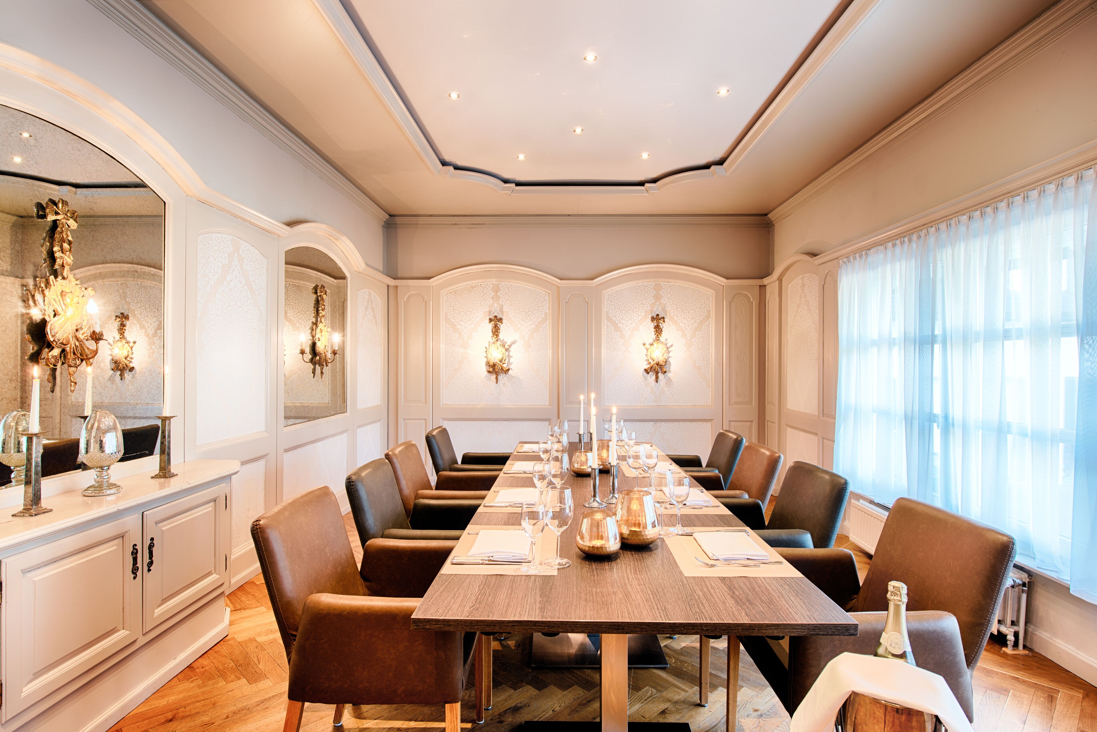 Un comedor elegante con una larga mesa, numerosas sillas, candelabros y un amplio ventanal con cortinas translúcidas.