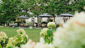 An outdoor seating area with tables and umbrellas, surrounded by lush greenery and white flowers. - Hotel Schloss Lehen (Bad Friedrichshall)