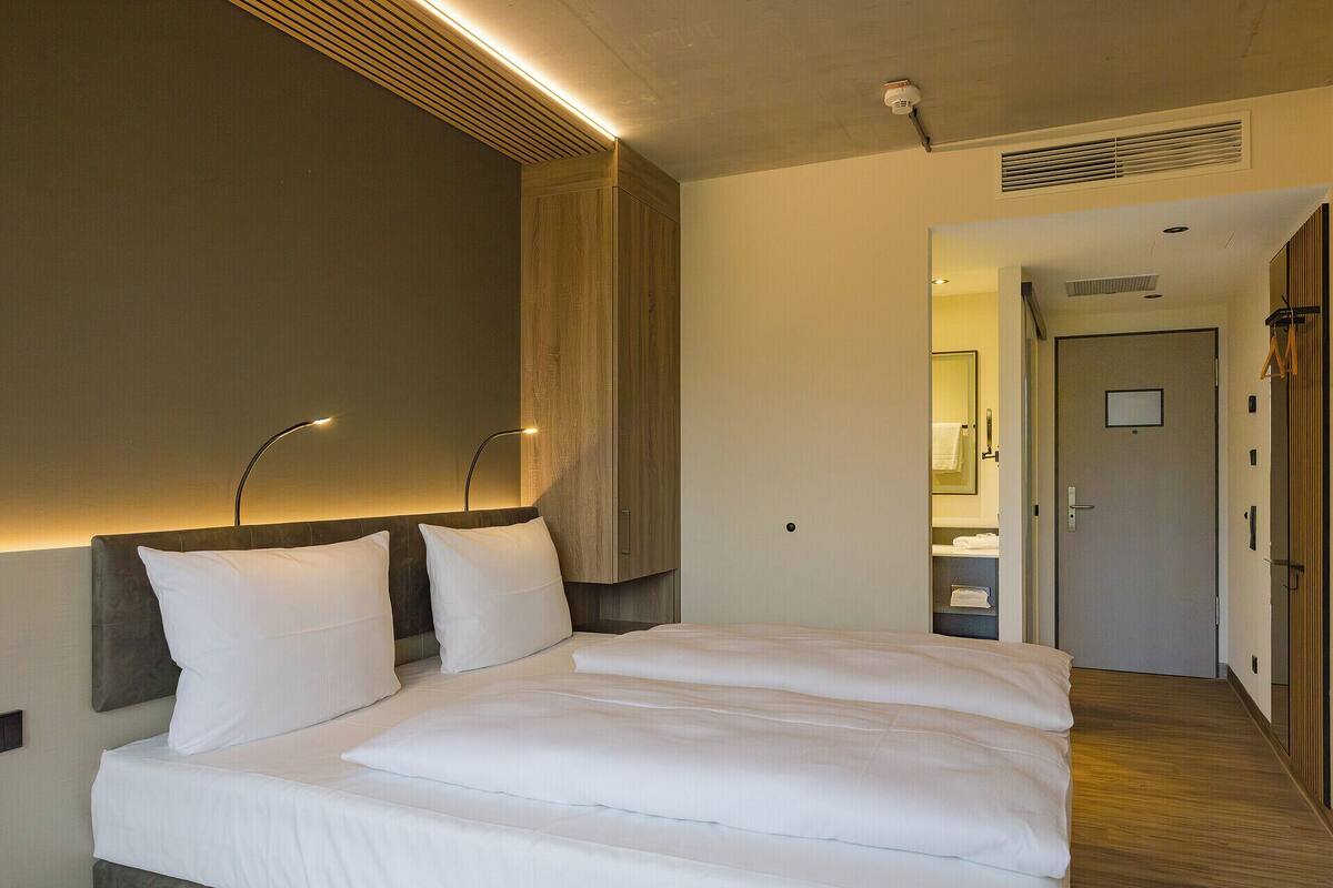 Habitació d'hotel amb un llit gran, un panell de capçalera de fusta, una taula de nit i una estantalla. Hi ha una porta que condueix a un bany i un passadís.