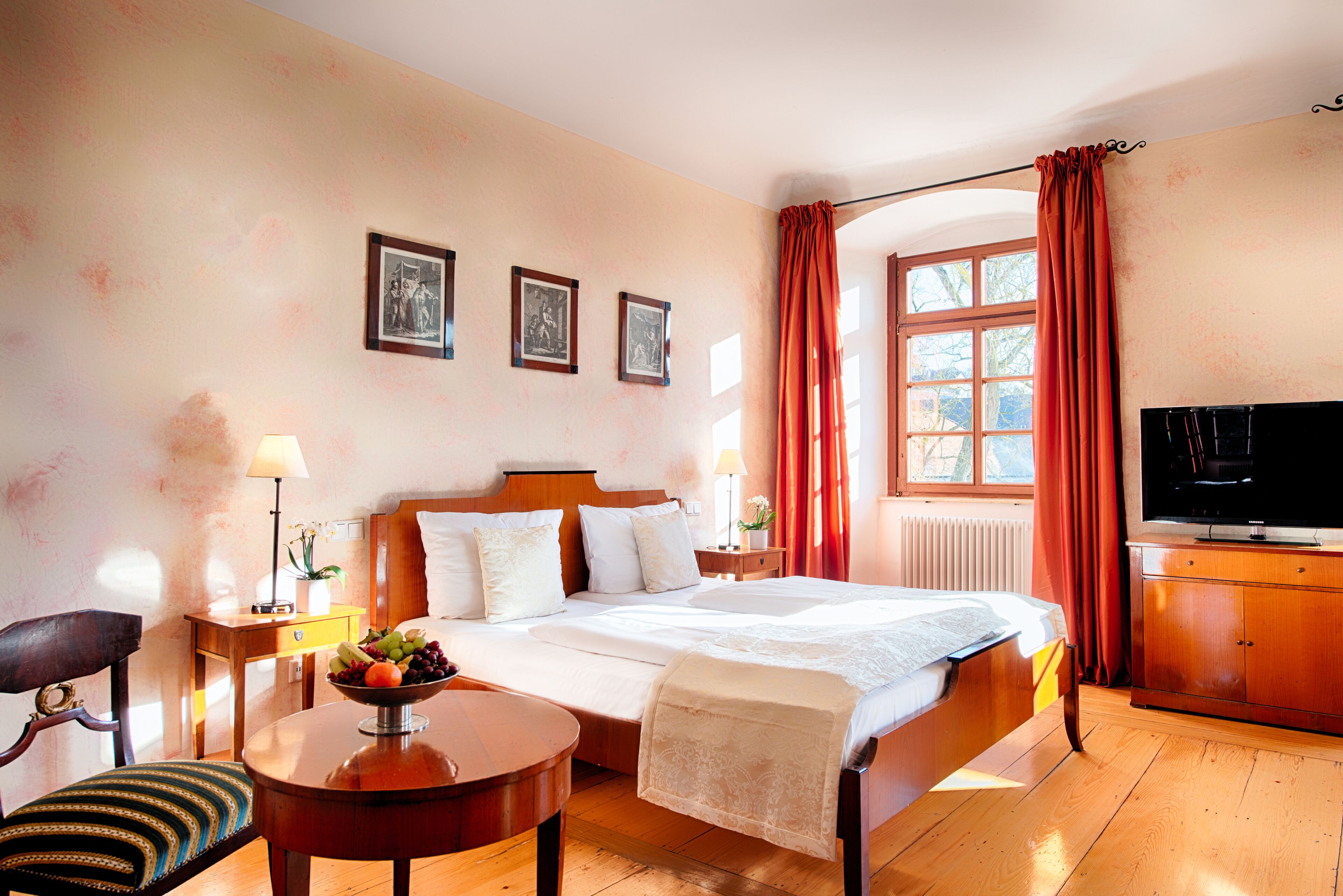 Ein Hotelzimmer mit einem großen Bett, einem Fernseher, einer Holzkommode, einem kleinen Tisch mit Obstschale, einem Sessel und einem Fenster mit Vorhängen.
