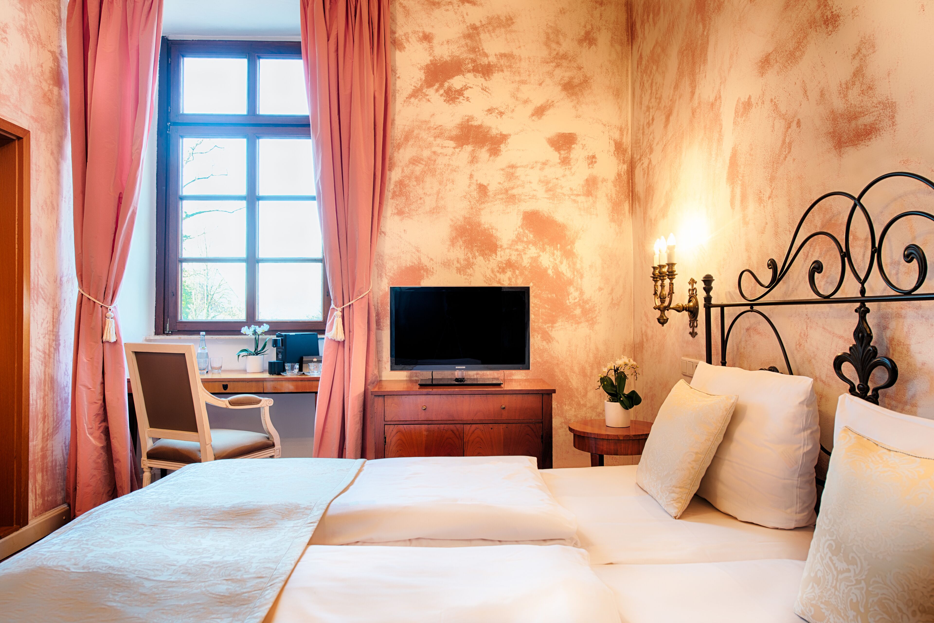 A hotel room with a bed, a desk, a television, and a wall-mounted lamp.