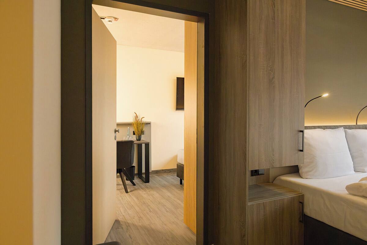Una habitació d'hotel moderna amb terra de fusta, un llit amb roba blanca, una petita taula de nit i una estantària.