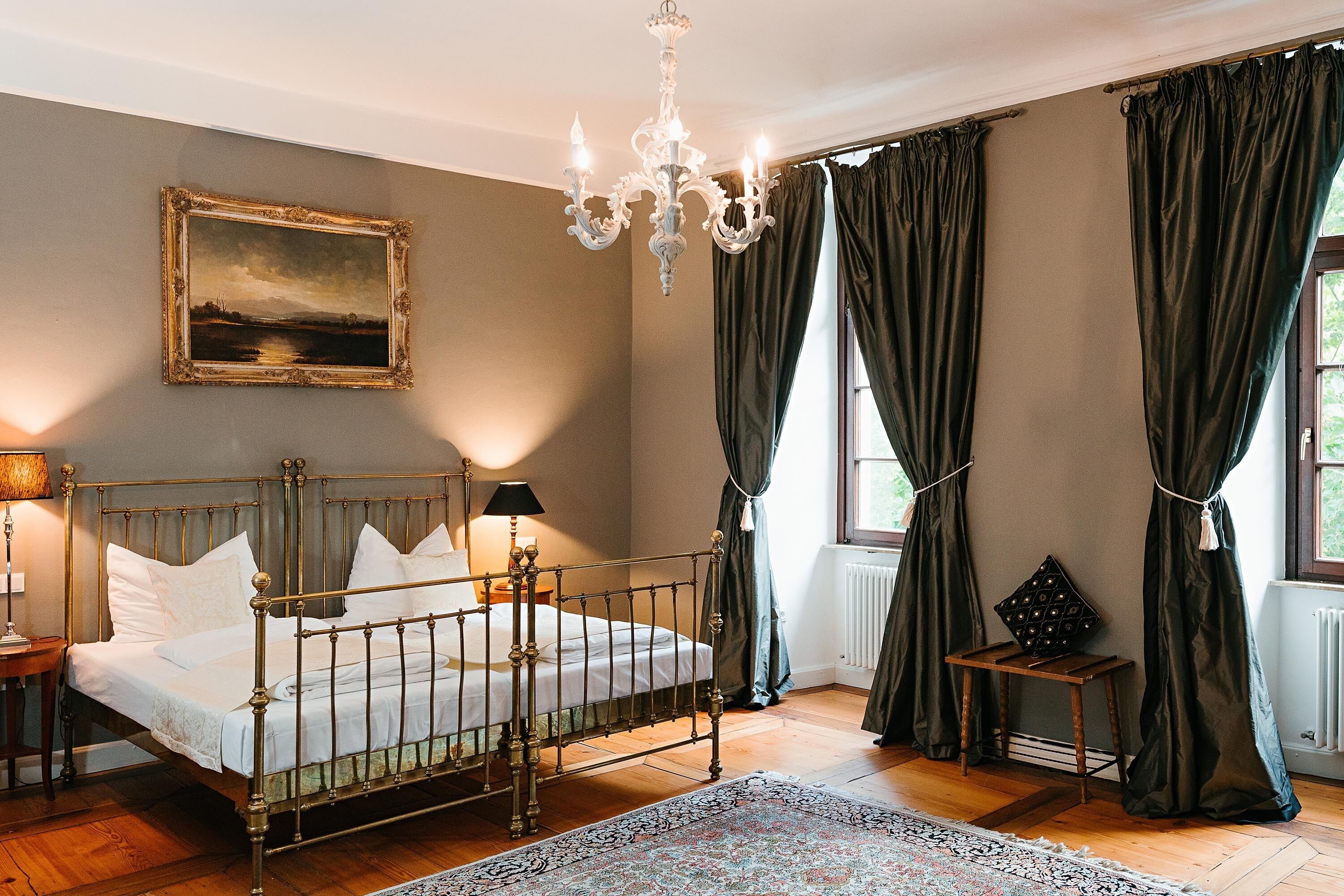 A bedroom with a metal bed frame, a chandelier, a painting, and a view of the outdoors through a window with curtains.