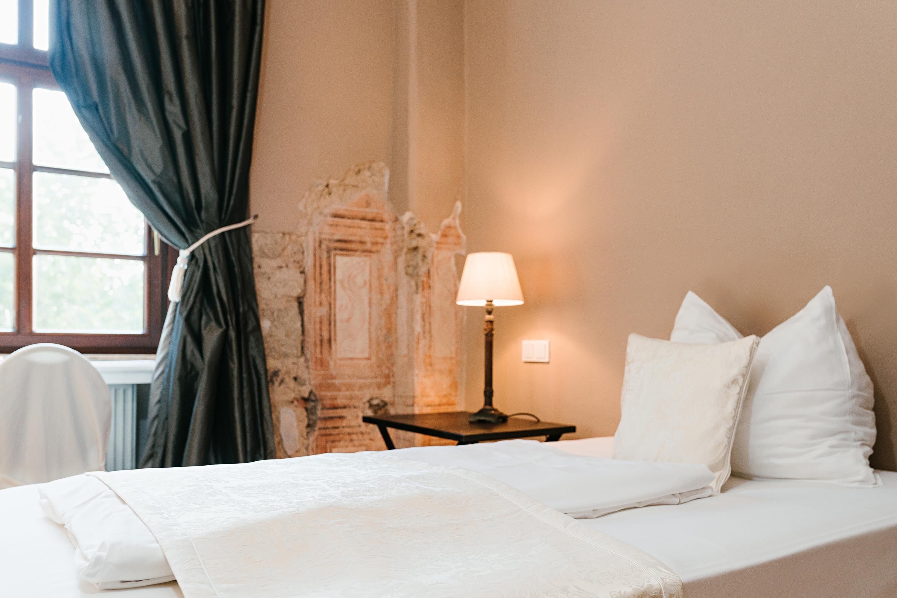 Un lit bien fait, avec du linge de lit blanc, une lampe de chevet et une chaise dans une pièce dotée d’une fenêtre, d’un mur orné d’un motif décoratif et d’un radiateur.