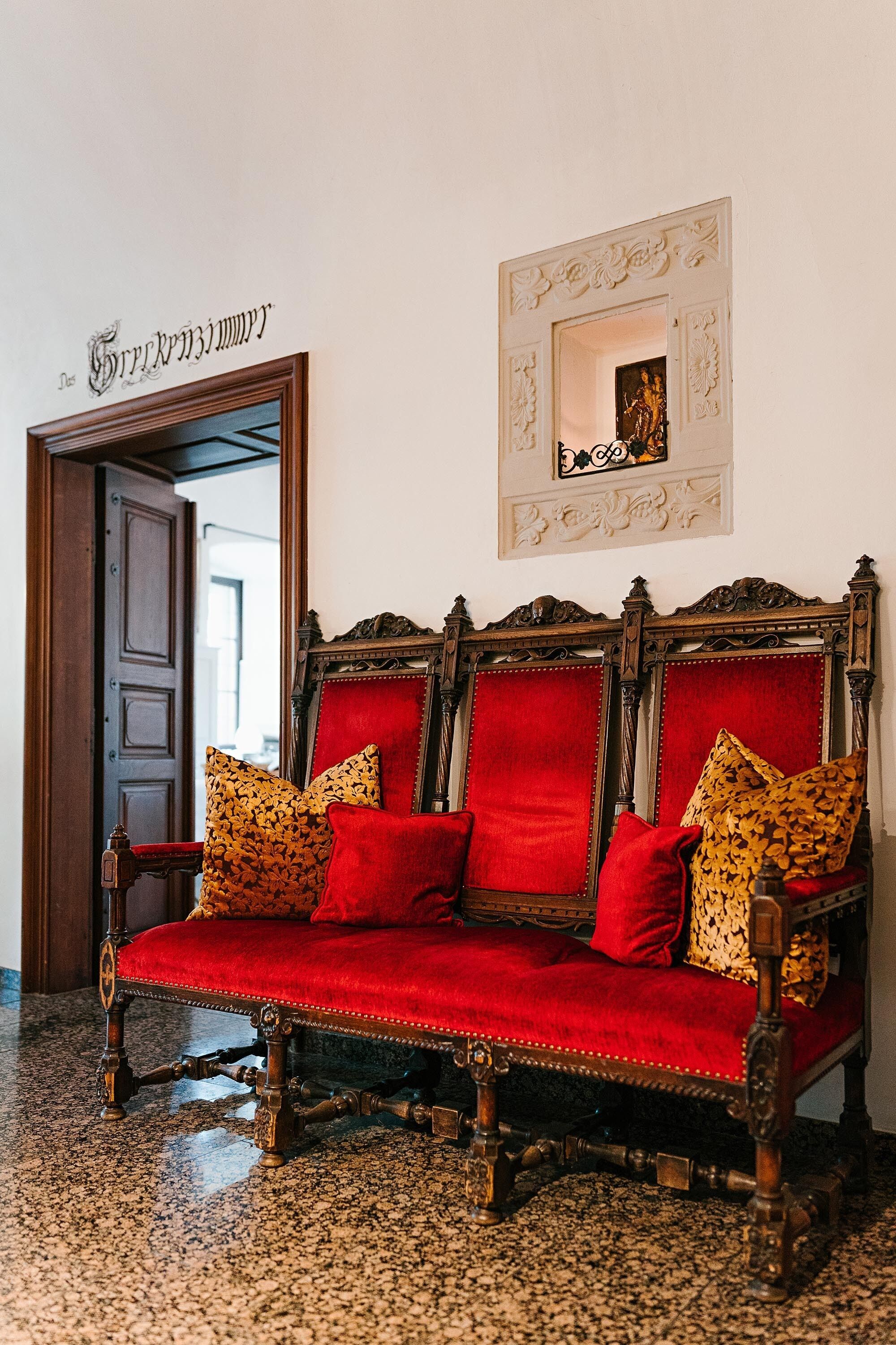 Un canapé traditionnel en bois, recouvert d’un tissu rouge et agrémenté de coussins à motifs, est placé dans un couloir où l’on aperçoit une porte en bois et un tableau encadré au mur.
