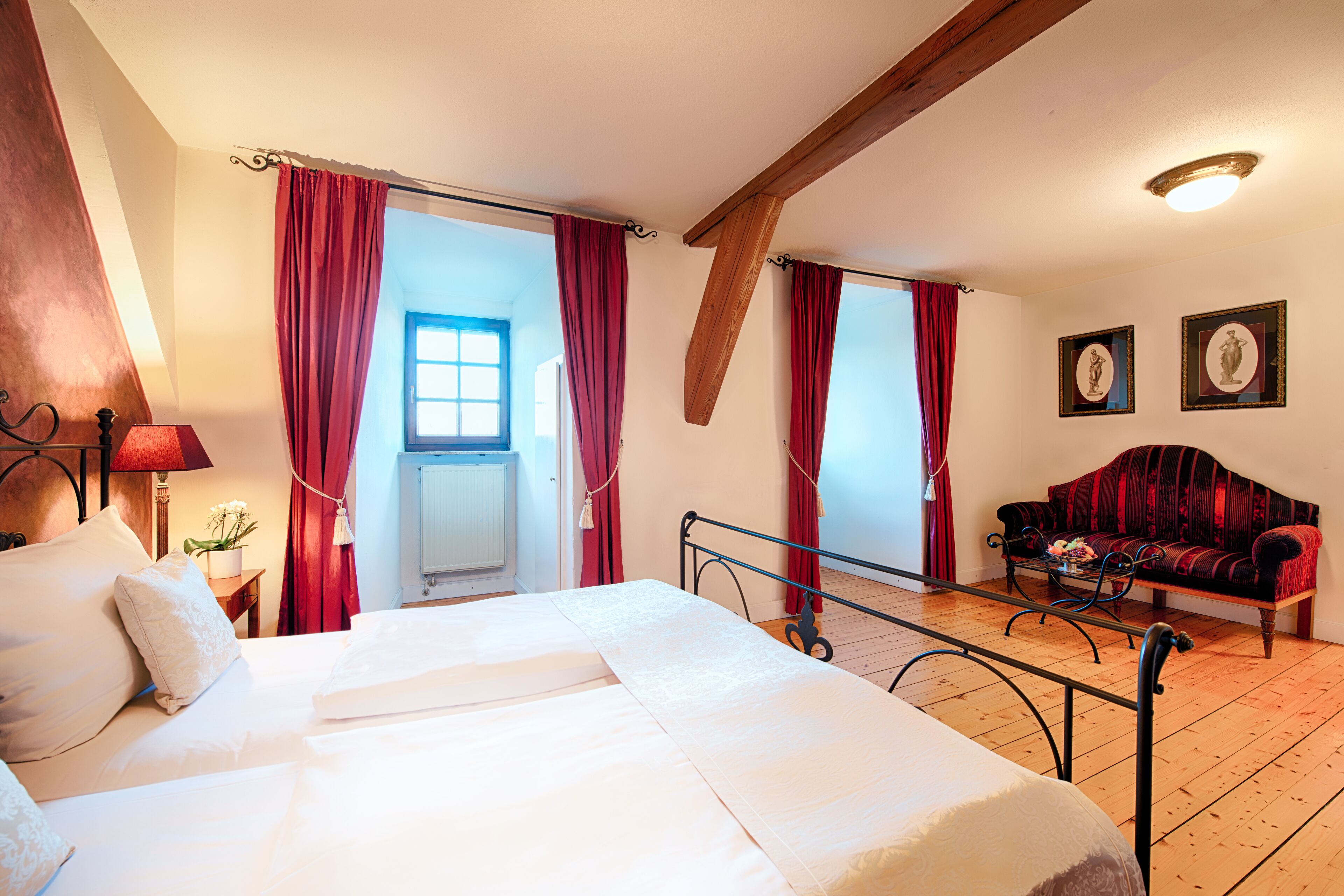 A bedroom with a bed, a wooden ceiling, a window, a red sofa, and a framed picture on the wall.