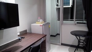 A small, modern room with a wooden desk, a flat-screen TV, a mini-fridge, a small table, and a sliding glass door leading to a balcony. - Pho Place (Bangkok)