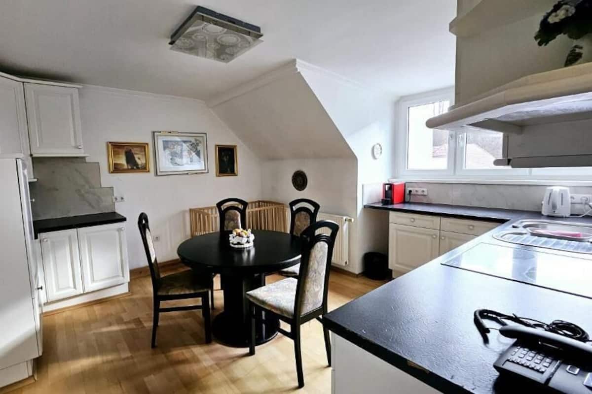 Een keuken met witte kastjes, een eettafel met stoelen, een zwart aanrecht en een raam.