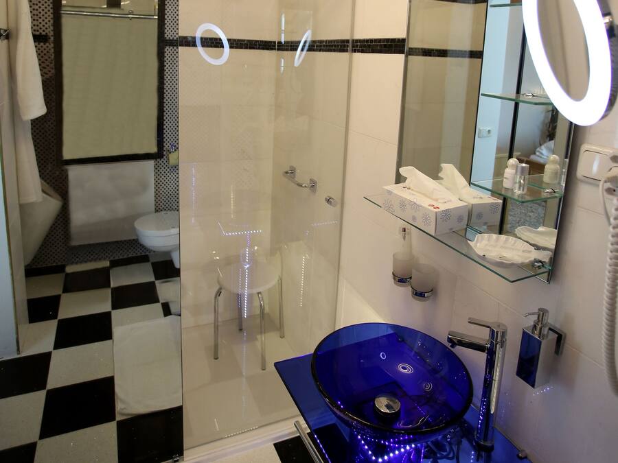 Un baño moderno con ducha envuelta en cristal, un lavamanos de vidrio azul y un espejo grande con luz redonda.