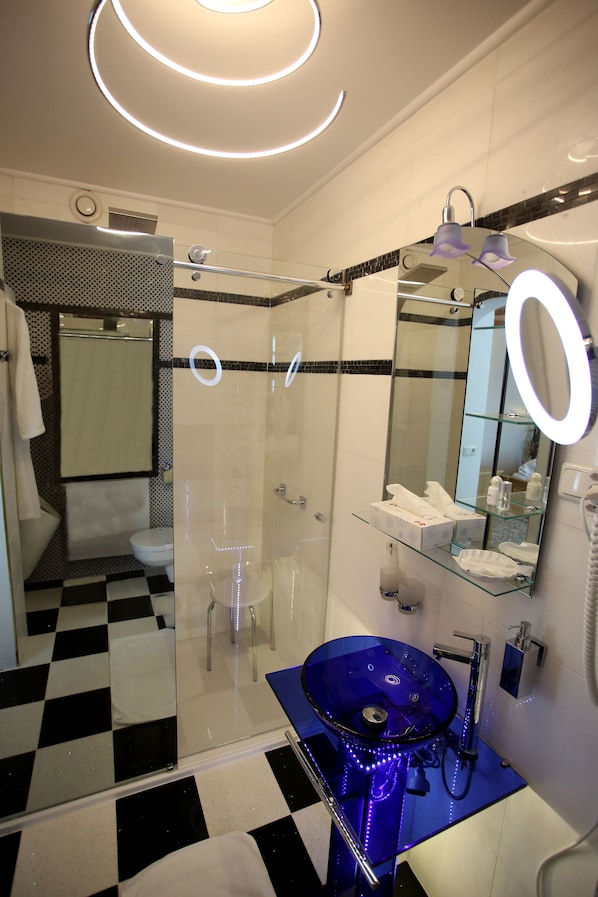 Un baño moderno con ducha envuelta en cristal, un lavamanos de vidrio azul y un espejo grande con luz redonda.