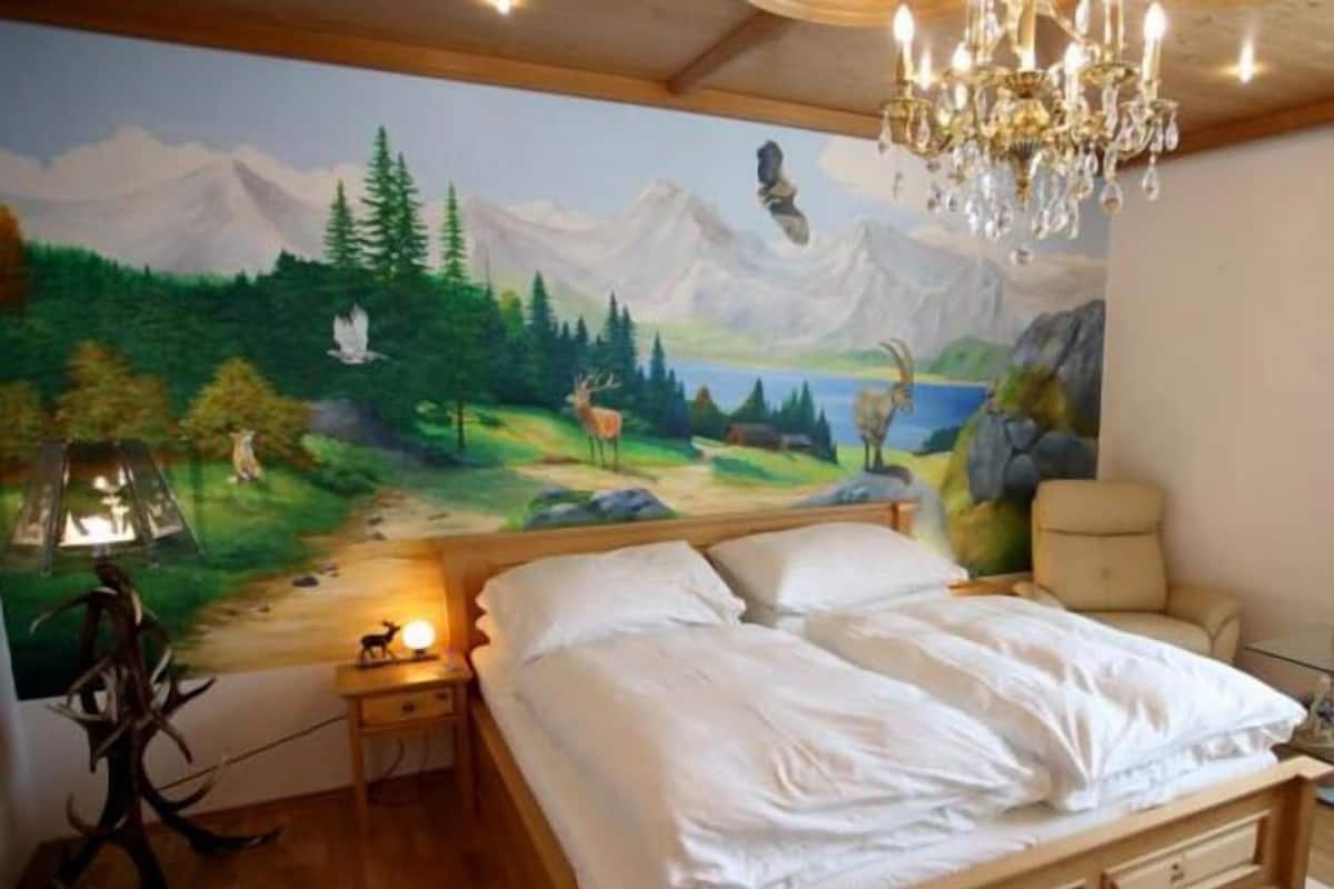Een slaapkamer met een houten bed, een nachtkastje met een lamp, een muurschildering van een boslandschap en een kroonluchter.