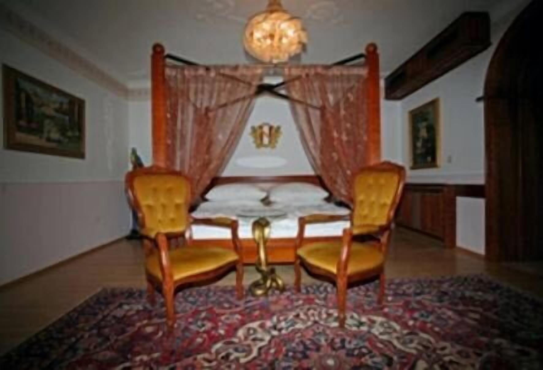 A room with a four-poster bed, two upholstered chairs, a patterned rug, and a chandelier.