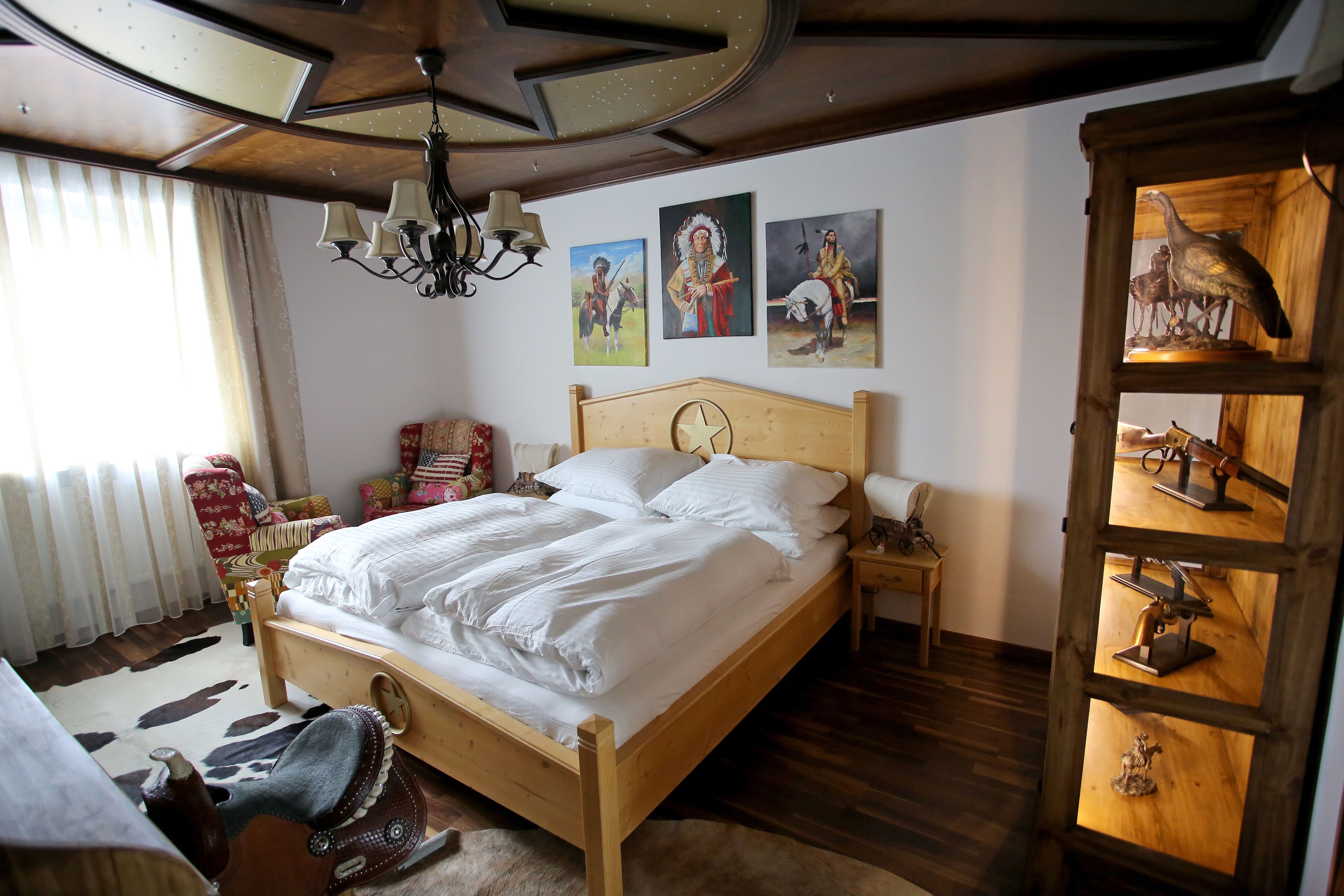 Una cama bien hecha con ropa blanca, cabecera de madera y mesita de noche con lámpara. Hay tres cuadros colgados en la pared que representan figuras históricas.