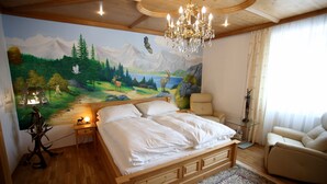 Une chambre à coucher avec un grand lit, un lustre, une fresque représentant une scène de forêt, une table de chevet en bois et une chaise.