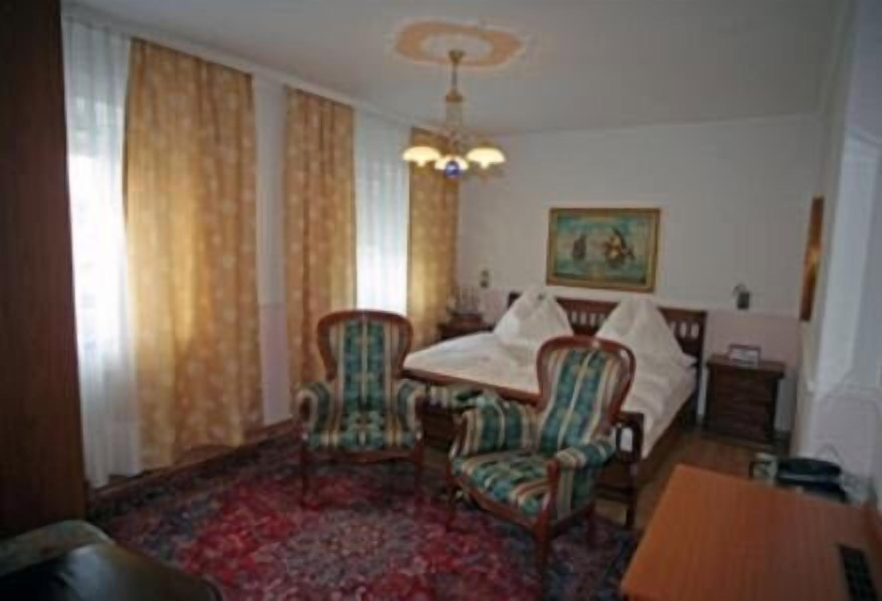 Een hotelkamer met een bed, twee fauteuils, een tafel, een kroonluchter en een schilderij aan de muur.