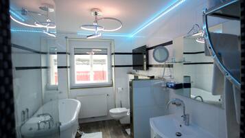 A modern bathroom with a bathtub, toilet, and sink. There is a window with a view of a shipping container outside.