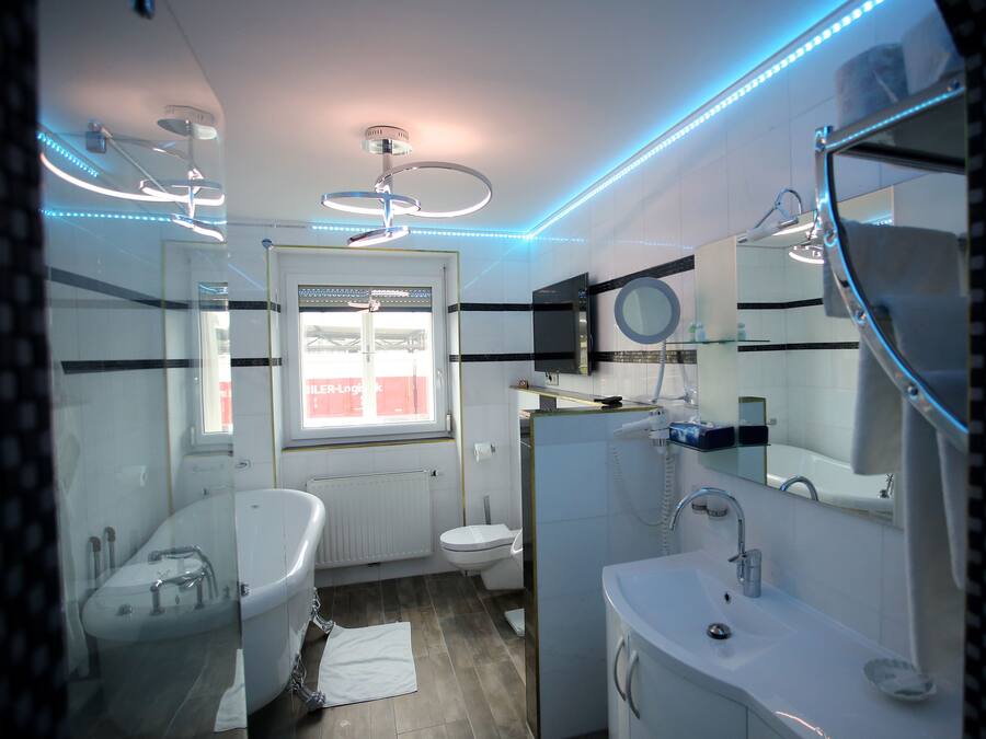 A modern bathroom with a bathtub, toilet, and sink. There is a window with a view of a shipping container outside.