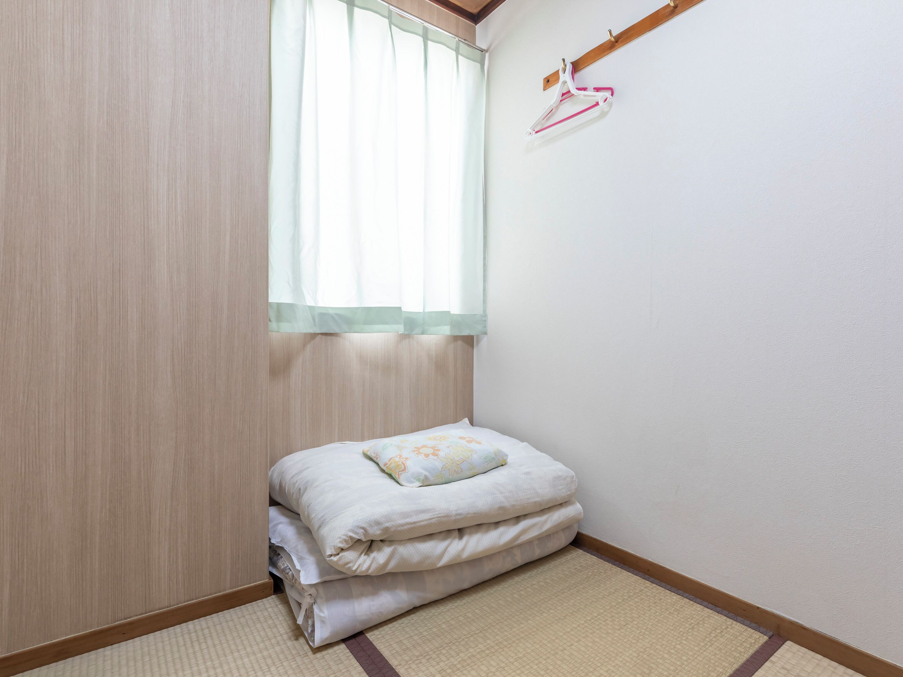 Ein kleines, ordentlich eingerichtetes Schlafzimmer mit einem Bett, einem Fenster mit transparenten Vorhängen und einem Holzkopfteil.