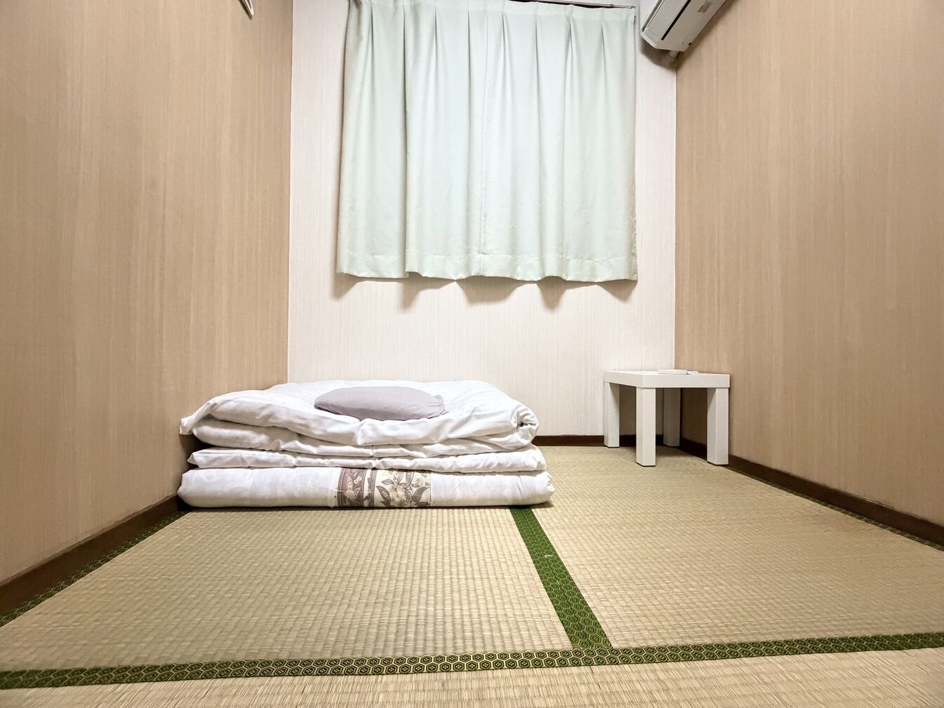A room with tatami flooring, a bed with pillows, a small table, and a window with a curtain.