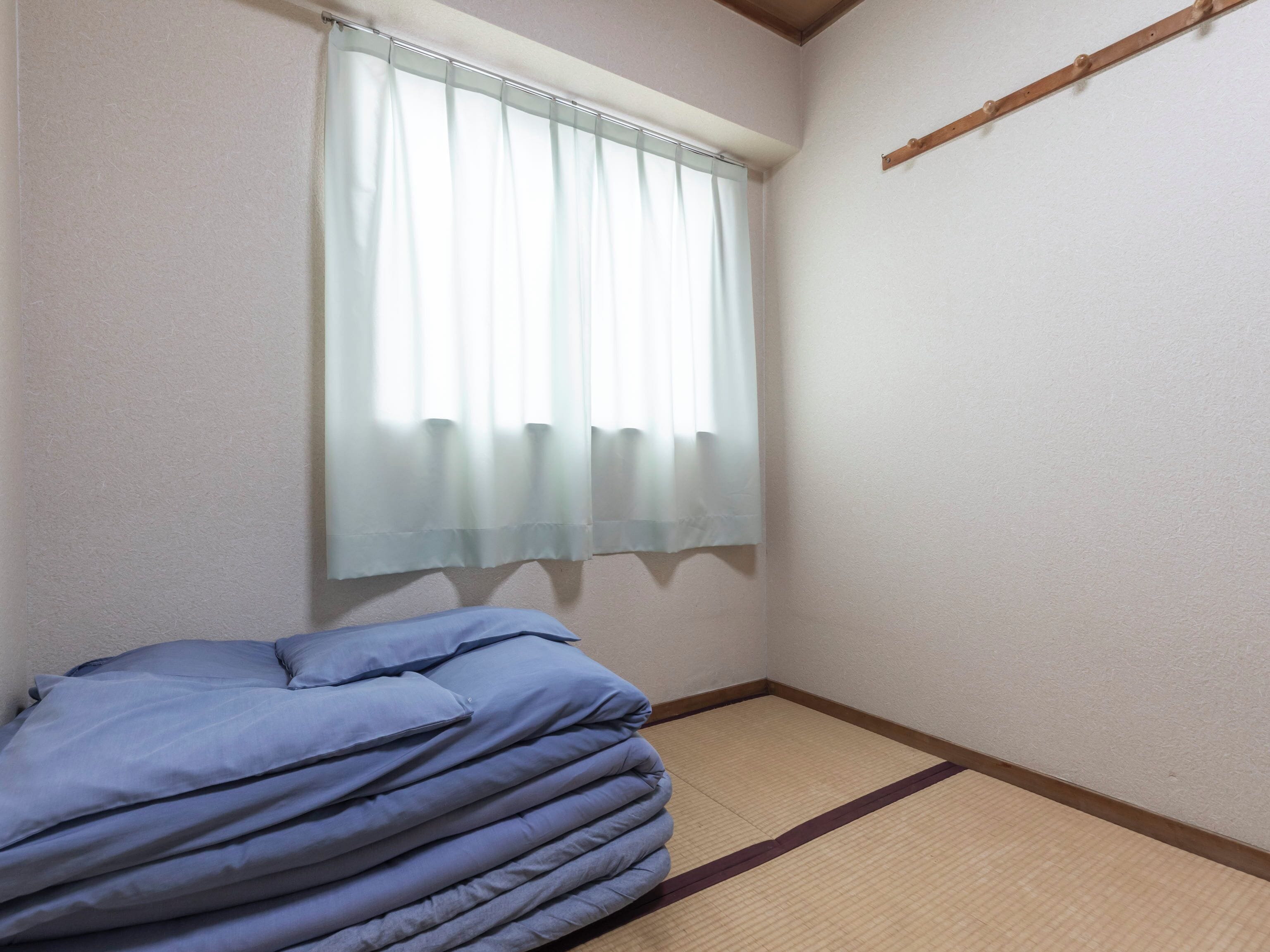 Une pièce avec un lit, une fenêtre ornée de rideaux légers, un sol recouvert de tatamis et une poutre en bois sur le mur.