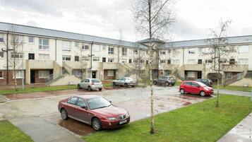 Un edificio residencial con varios pisos, un estacionamiento y una pequeña zona verde con árboles.