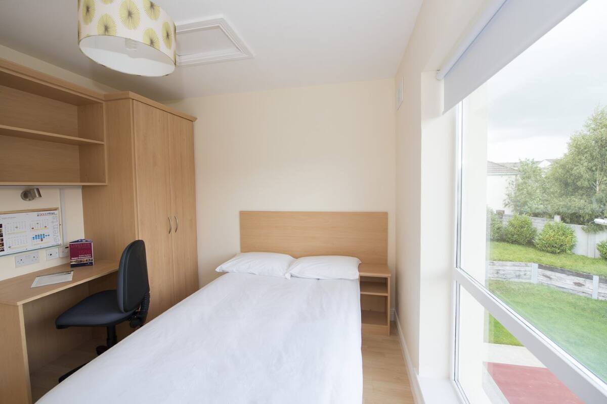 A bedroom with a bed, a desk, and a chair. There is a window with a view of a garden.