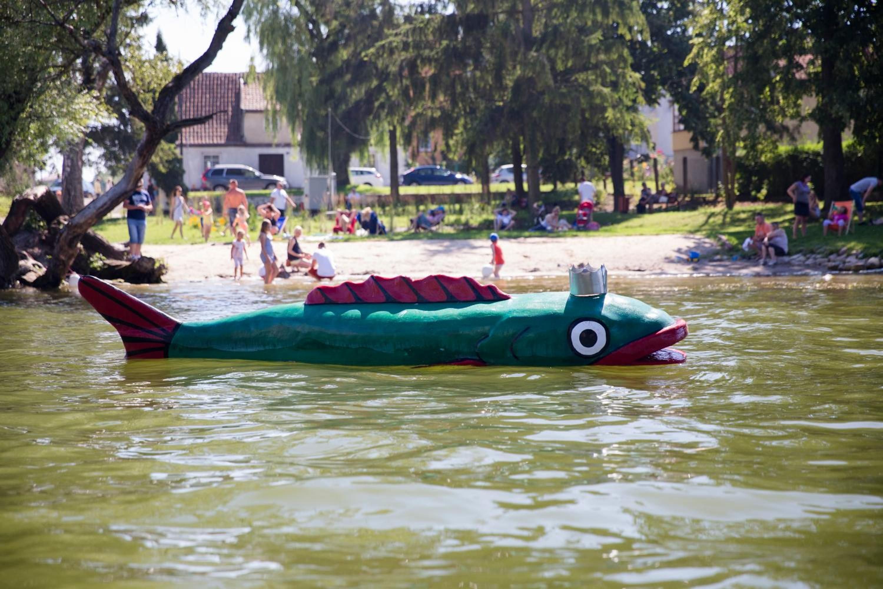 Un pez inflable verde en el agua, con gente de fondo.