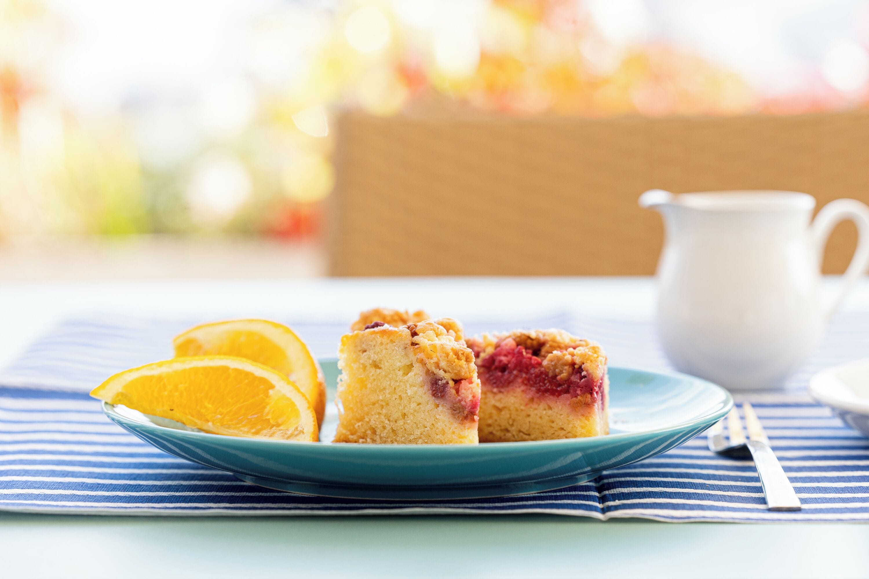A plate with two pieces of cake and a slice of orange on a blue plate, a white pitcher, and a striped napkin on a table.