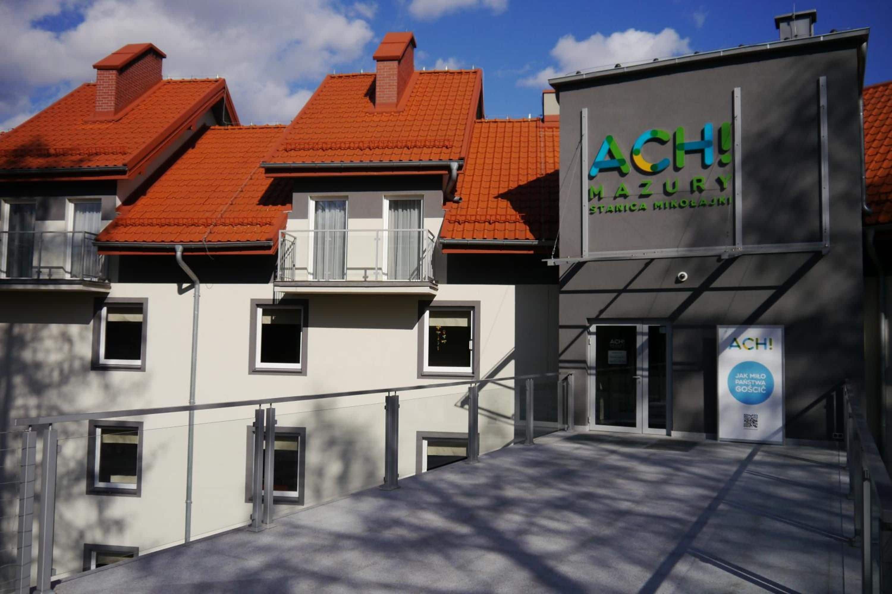A building with a red-tiled roof, a sign reading 'ACH Mazury,' and a modern entrance with glass doors.