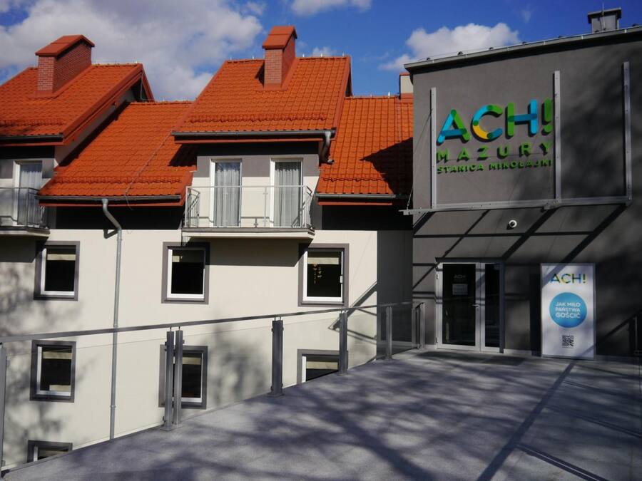 A building with a red-tiled roof, a sign reading 'ACH Mazury,' and a modern entrance with glass doors.