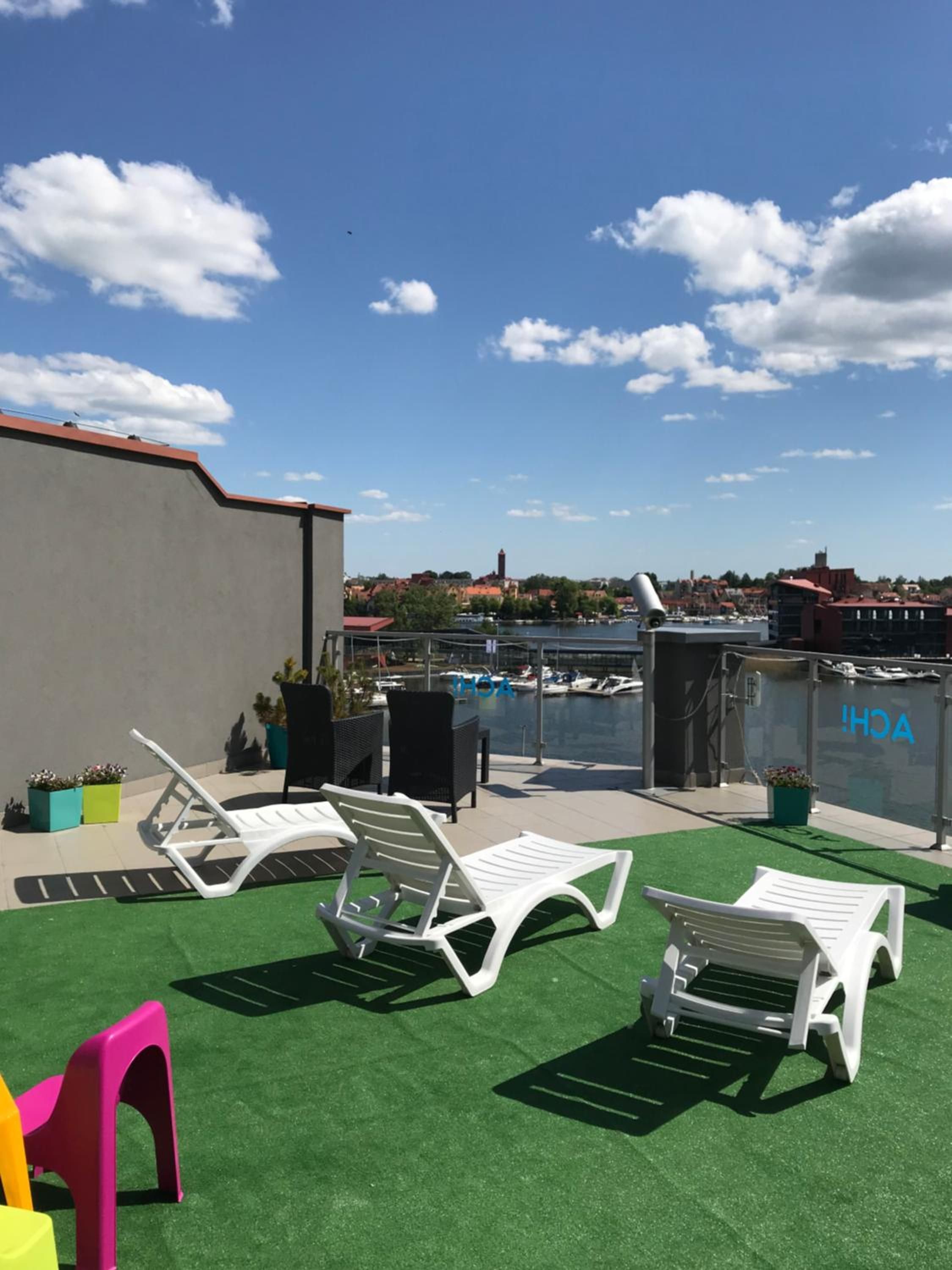 A rooftop terrace with white lounge chairs, a green artificial turf area, and a view of a marina with boats.