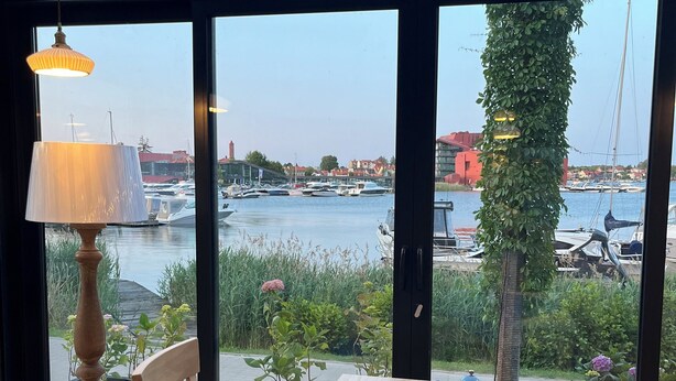 Una mesa de madera con mantel azul, dos sillas, una lámpara y vistas a barcos y vegetación a través de ventanas grandes.