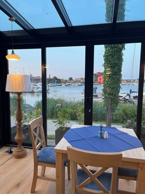 A wooden table with a blue tablecloth, two chairs, a lamp, and a view of boats and greenery through large windows. - Ach Mazury Stanica Mikołajki (Mikolajki)