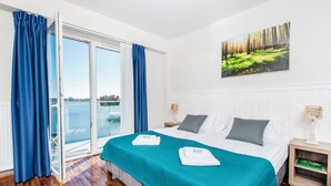 Habitación de hotel con una cama grande, vista al mar a través de una puerta corredera y un cuadro de un bosque en la pared.